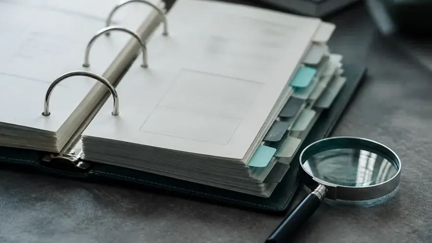 An open ring binder with tabbed dividers, a magnifying glass and someone taking notes